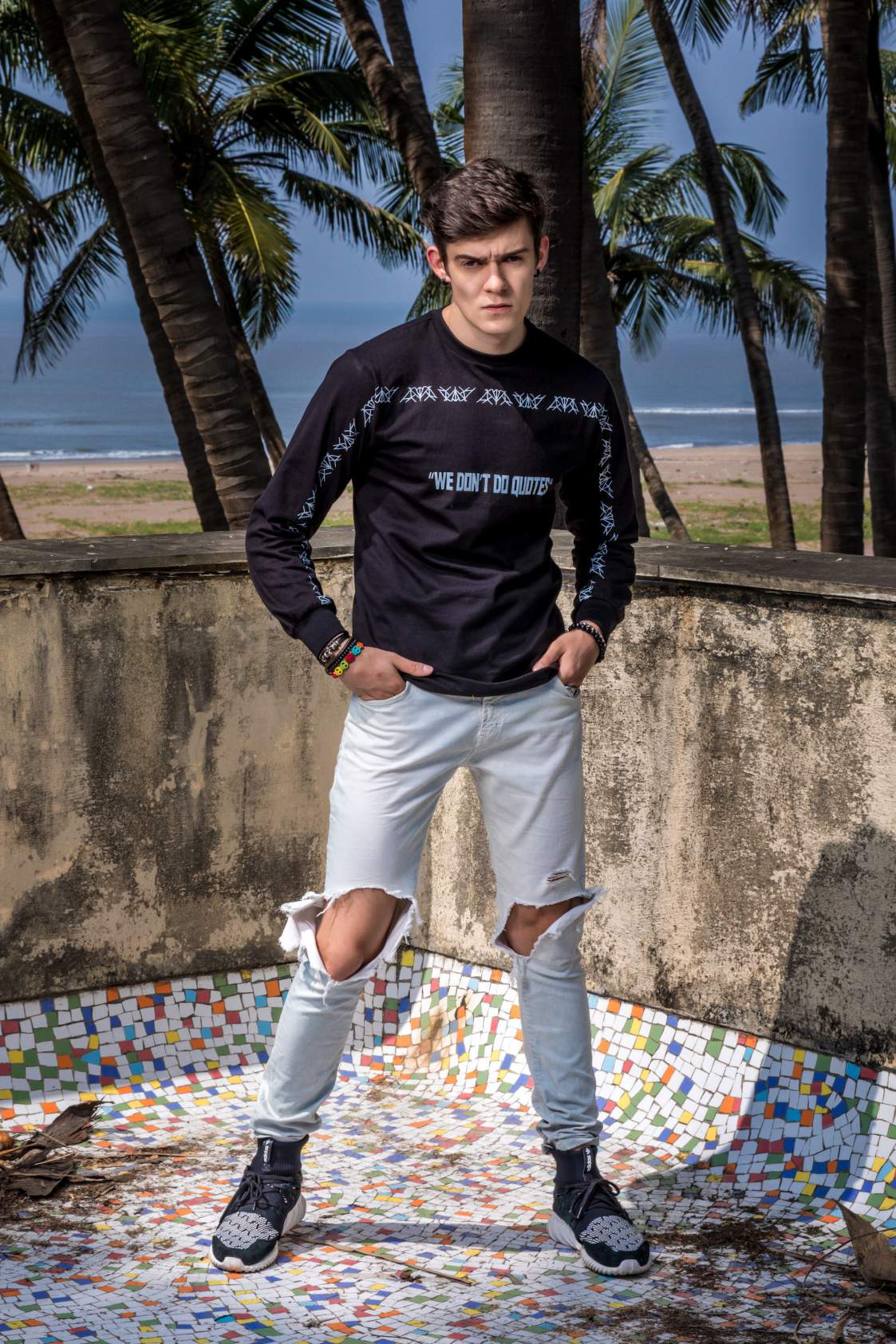 A boy wearing Black round neck full sleeve T-shirt paired with knee torn denim and black shoes is standing on the mosaic floor
