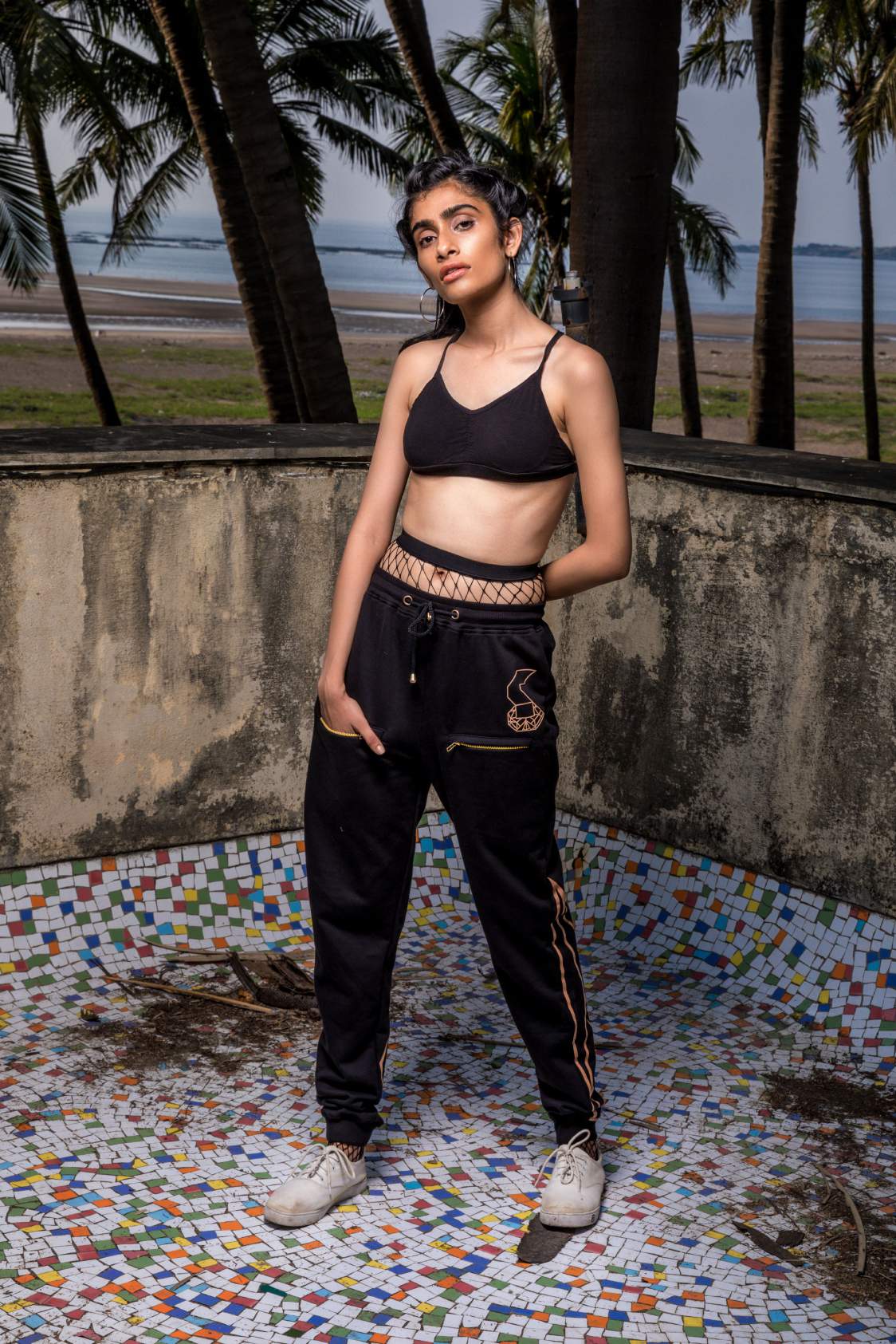 A girl wearing Black colored track pant featuring an elastic ankle and waist with adjustable drawstrings and front pockets with the logo right above the left pocket and geometric designs at the back of the track pant on the lower leg standing on mosaic titles.
