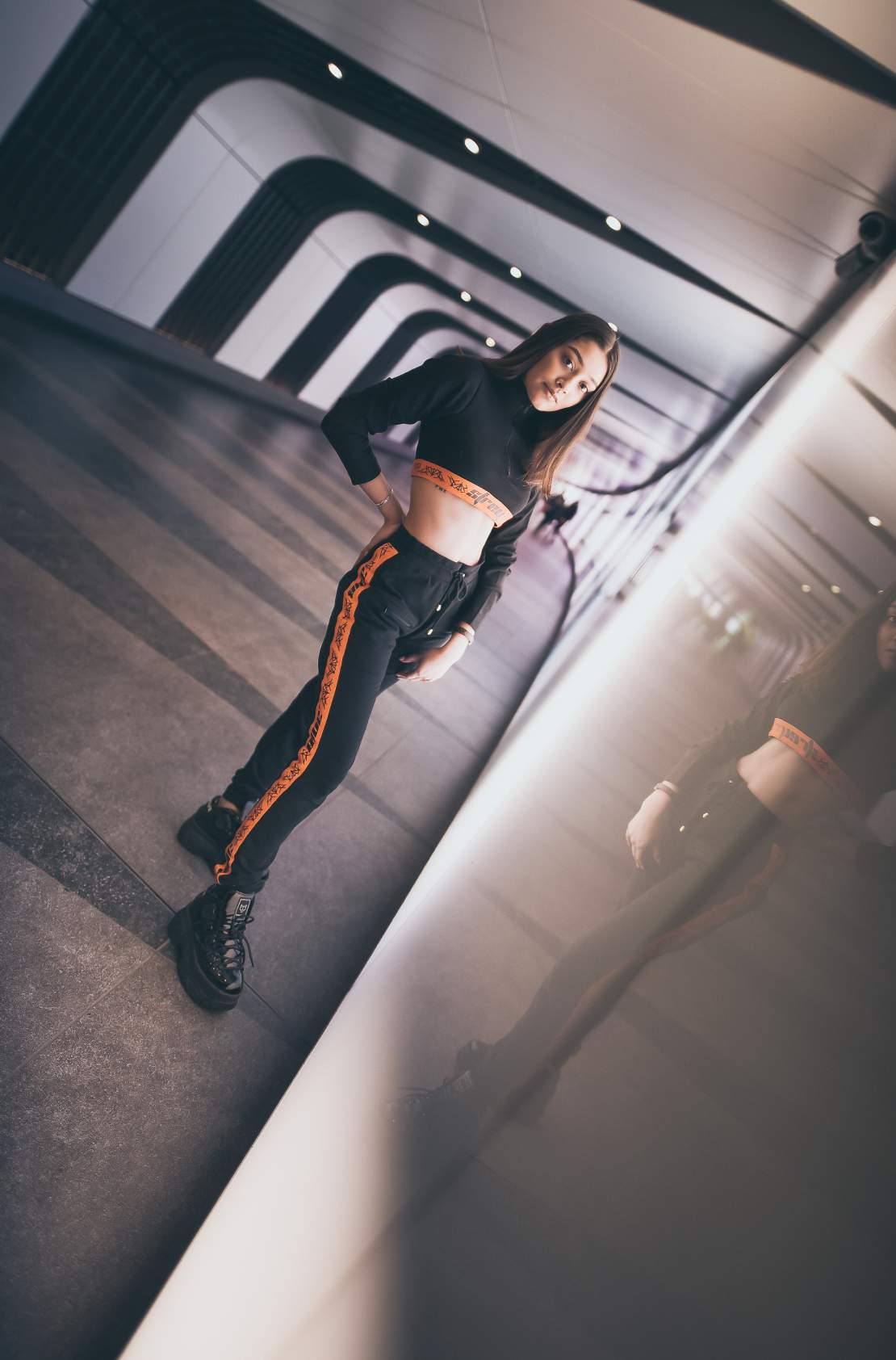 A girl wearing Black colored crop top with orange band at the bottom with a zip in the front and black and orange track pants with orange strips on the side of the track pant and high boots. Standing in a subway.