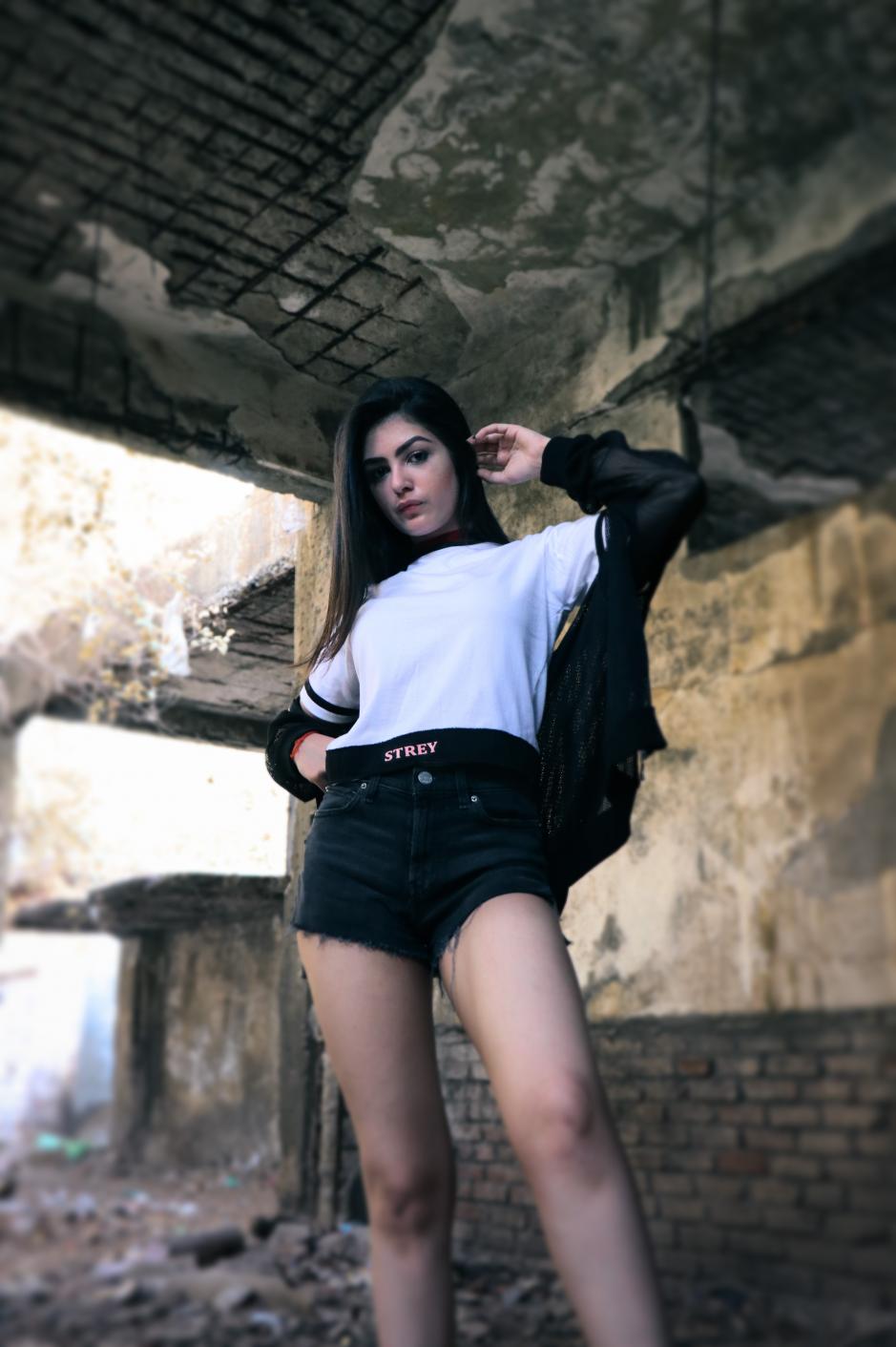 A girl wearing black and white round neck short sleeve regular fit T-shirt with black bands on neck sleeves and bottom of the T-shirt and black overcoat paired with shorts is standing in an under construction building.