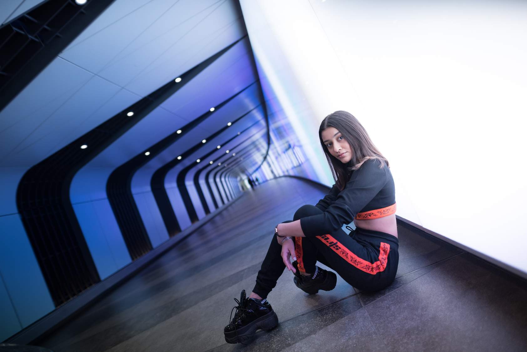 A girl wearing Black colored crop top with orange band at the bottom with a zip in the front and black and orange track pants with orange strips on the side of the track pant and high boots. Sitting in a subway.