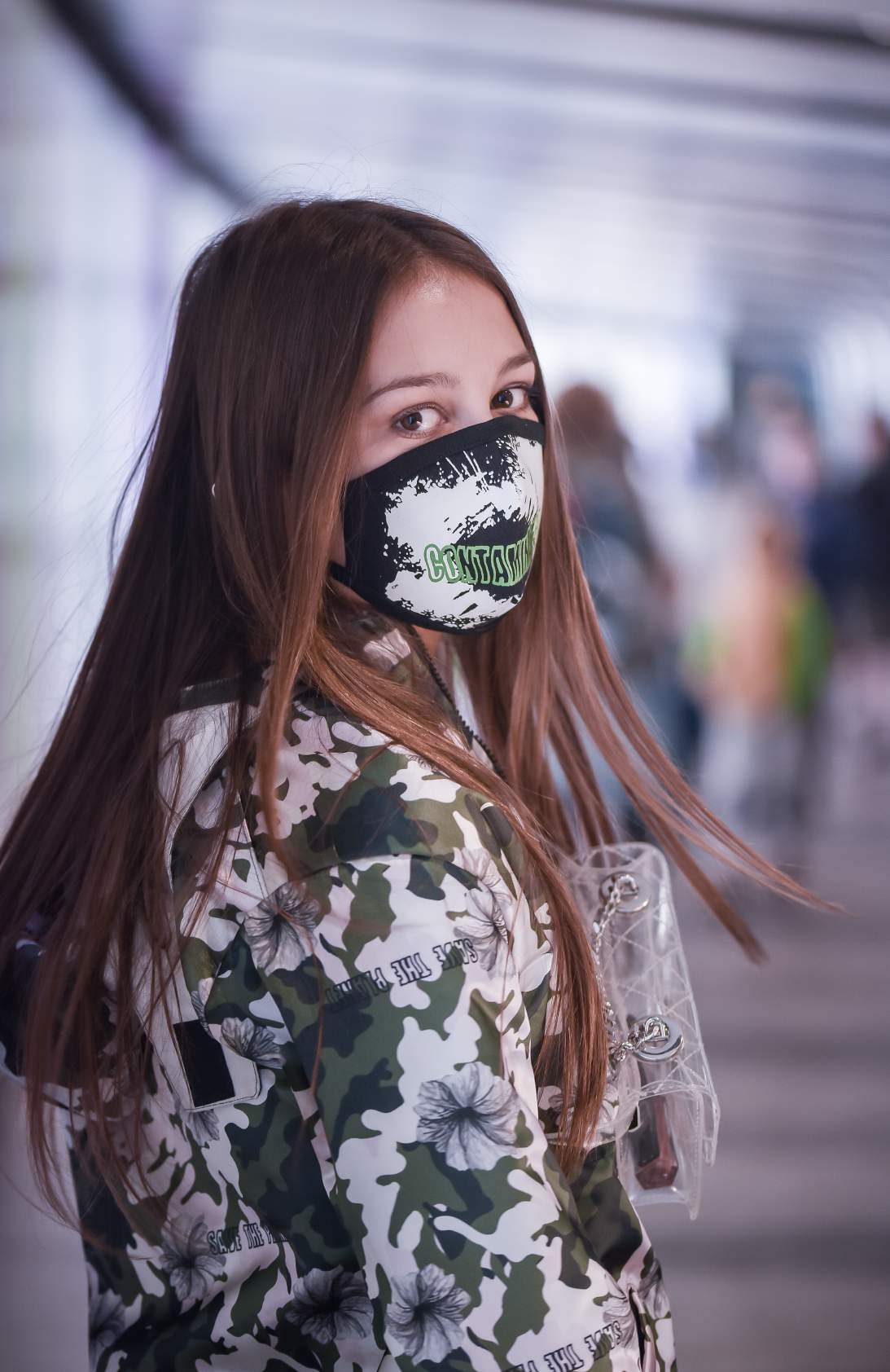 A girl wearing green and white coat and wearing a mask with a transparent sling around her neck is standing in the subway