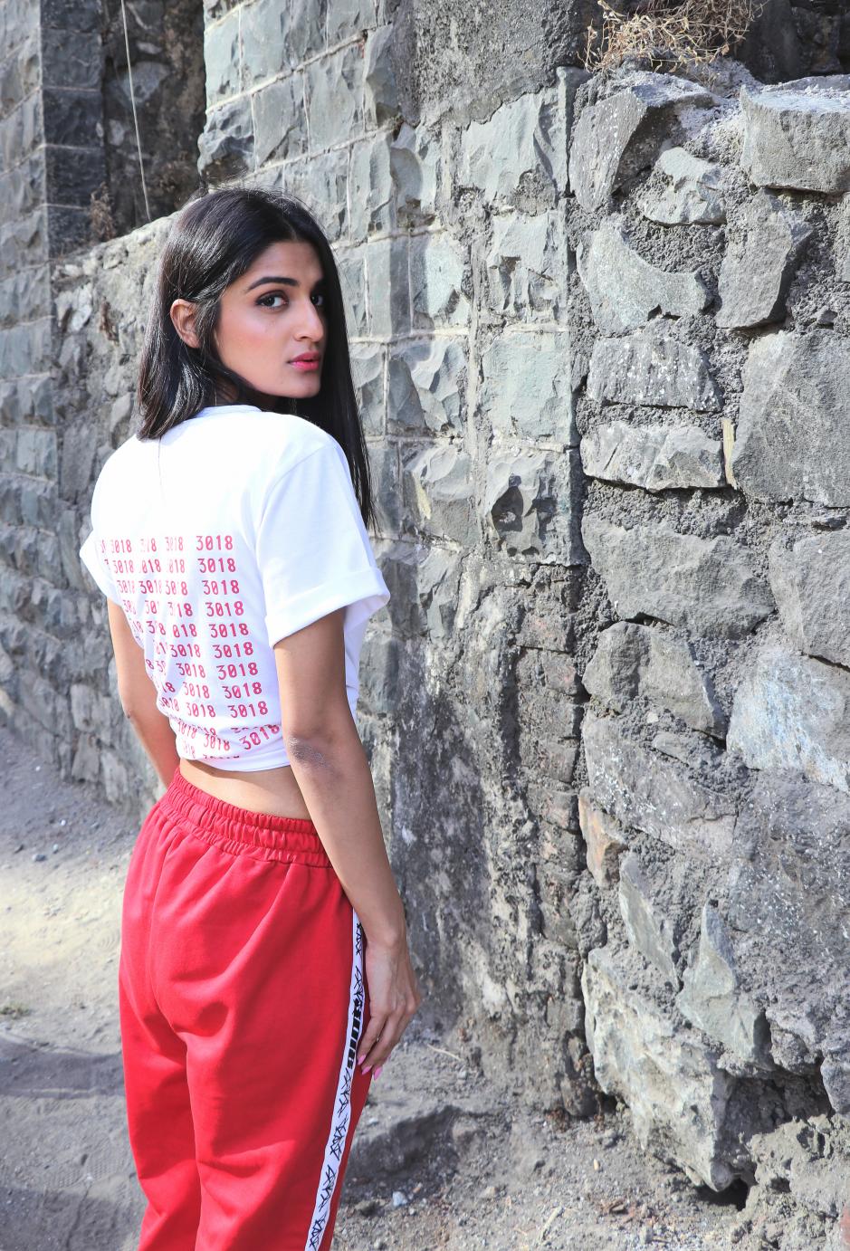 A girl wearing Red track pant with white strips on the sides and featuring elastic on the waist paired with a white T-shirt who is looking towards her side is standing in front of a rocked wall.