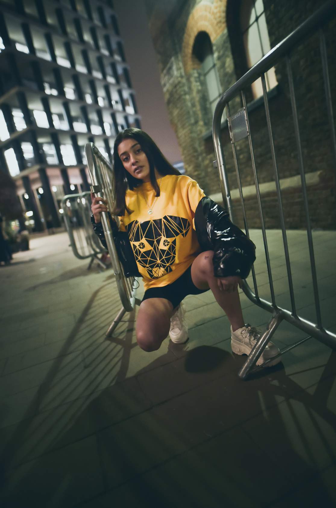 A girl wearing yellow round neck short sleeve regular fit T-shirt with dog printed on it paired with black shorts and shoes is sitting holding the gate.