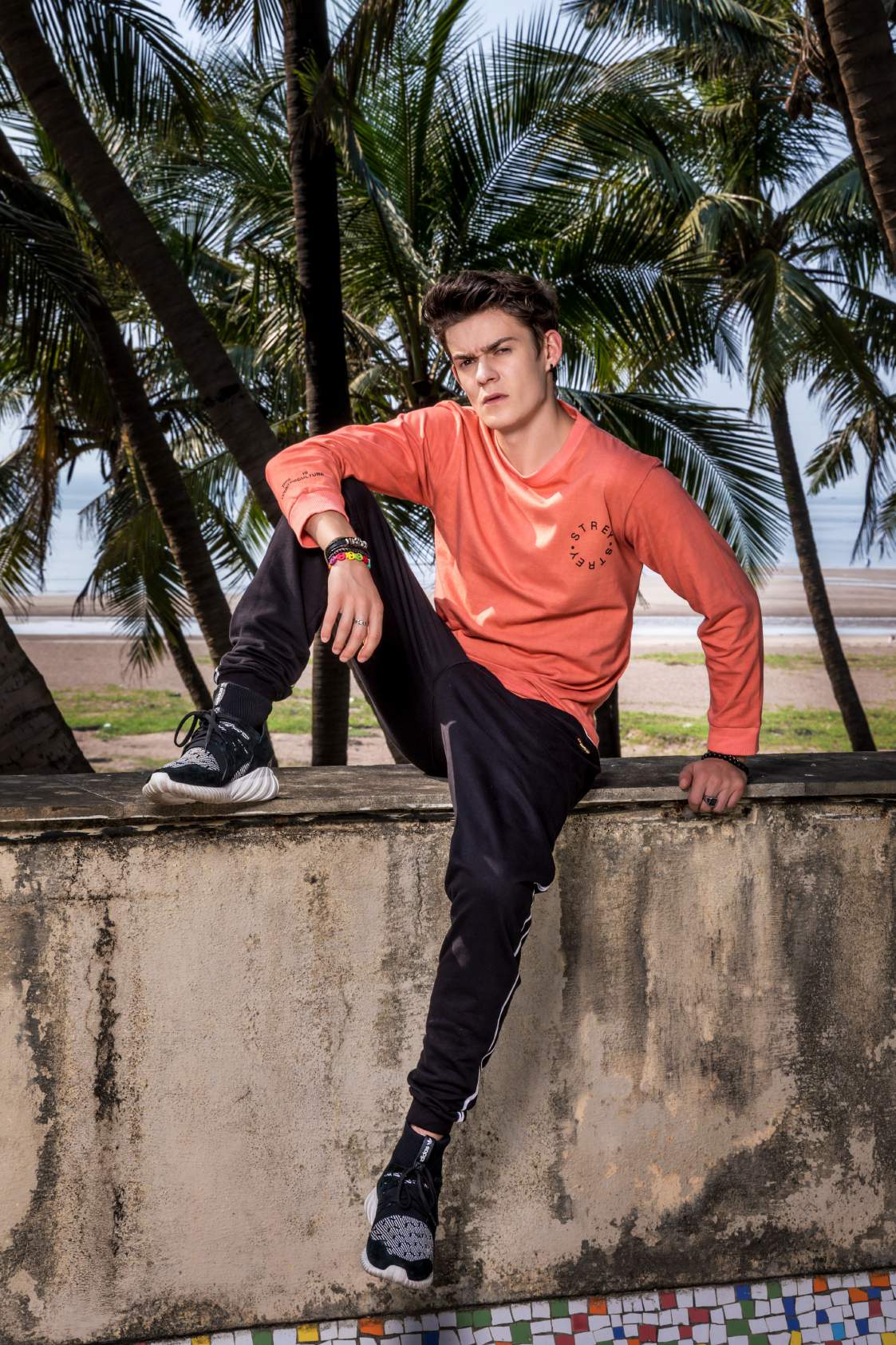 A boy Wearing pink full sleeved round neck T-shirt paired with black track pant and shoes is sitting on a wall.