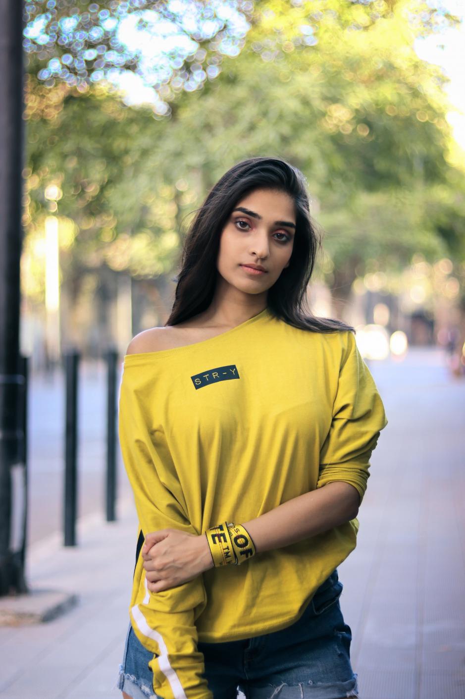 A girl wearing yellow round neck full sleeves T-shirt with white strips on the sleeves and str-y written on it is standing on a footpath