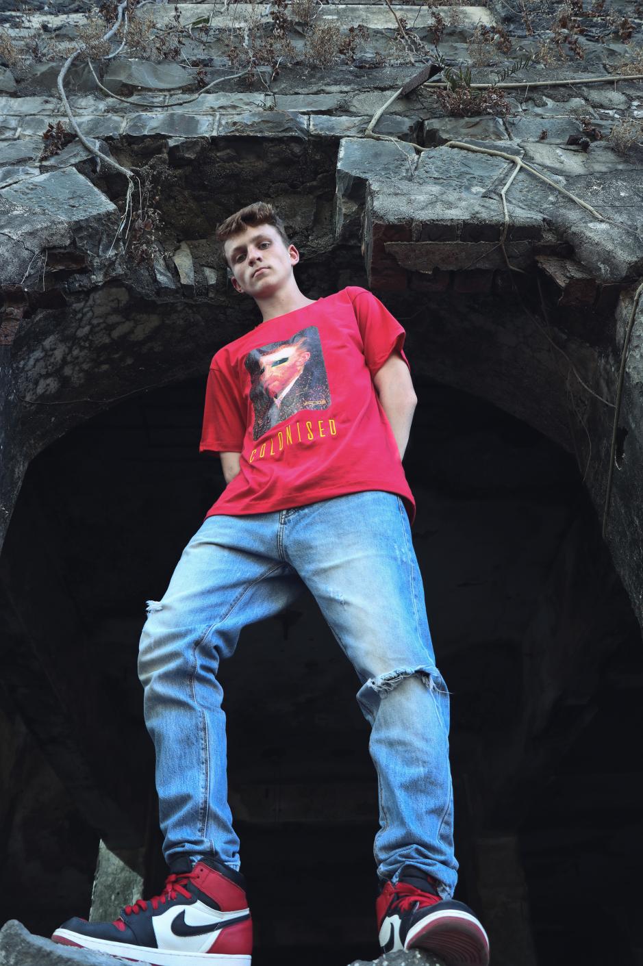 A boy wearing red short sleeve round neck T-shirt paired with a blue knee torn denim and red black shoes is standing on top a rock wall.