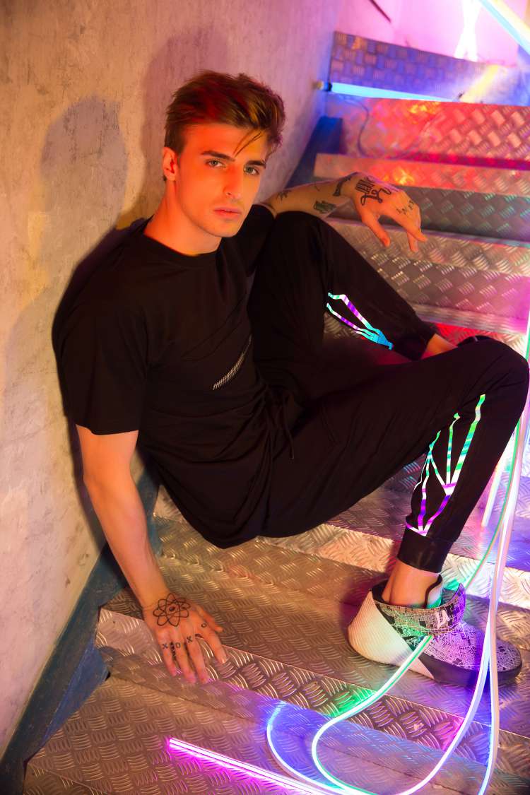 A boy wearing Black regular fit short sleeve round neck T-shirt with a lots of f printed and ripped in the center paired with black track pant and shoes is sitting on the staircase with colored led strip.