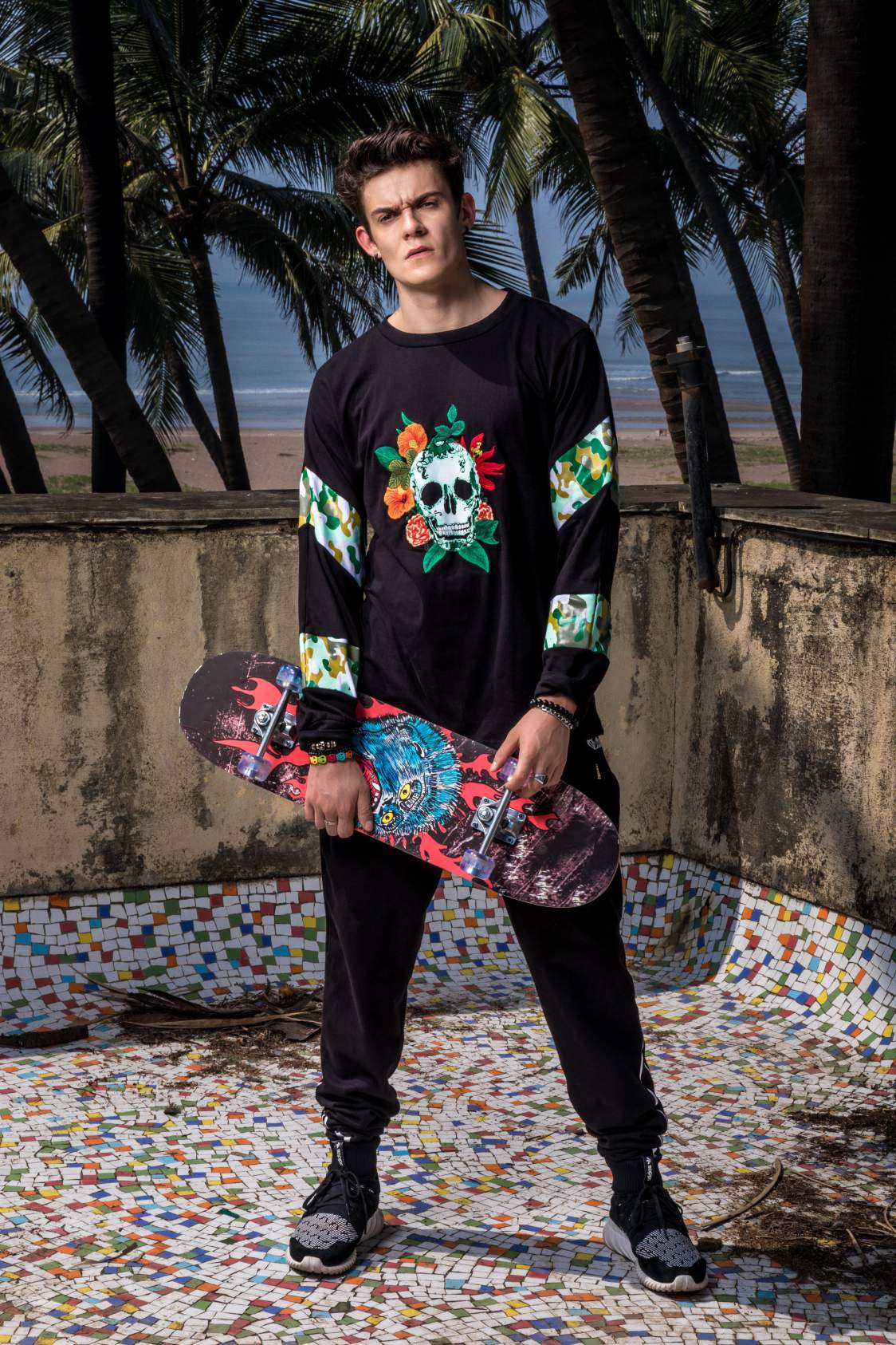 A boy wearing a black round neck full sleeve T-shirt with elastic on the wrist and a Skull design in the center of the T-shirt with 2 printed strips on each sleeve and black track pant is standing and holding the skateboard with his right hand. 