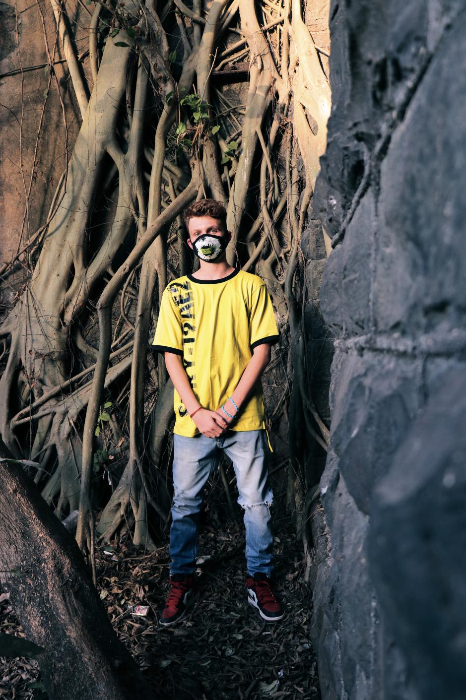 A boy wearing yellow and black round neck short sleeves regular fit T-shirt with distorted STRY-19AE2 printed on it with blue denim red shoes and mask is standing under a tree