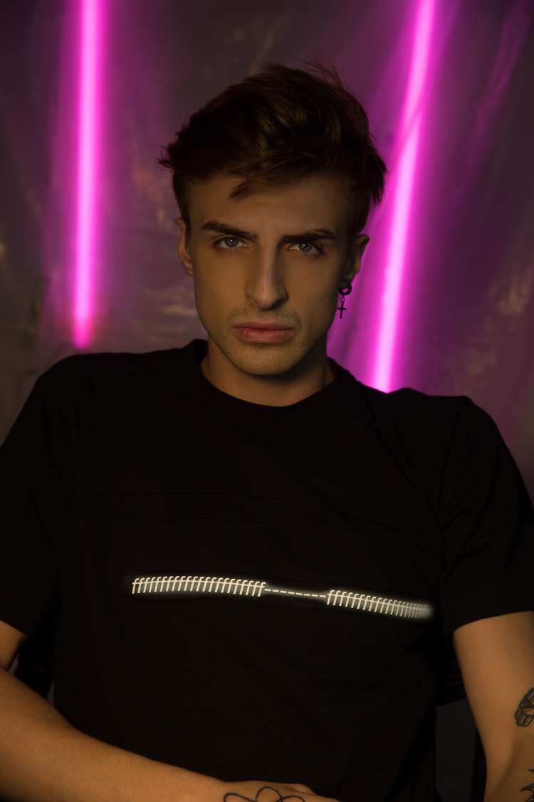 A Boy wearing black regular fit short sleeve round neck T-shirt with a lots of reflective f printed and ripped in the center with a purple light in the background.