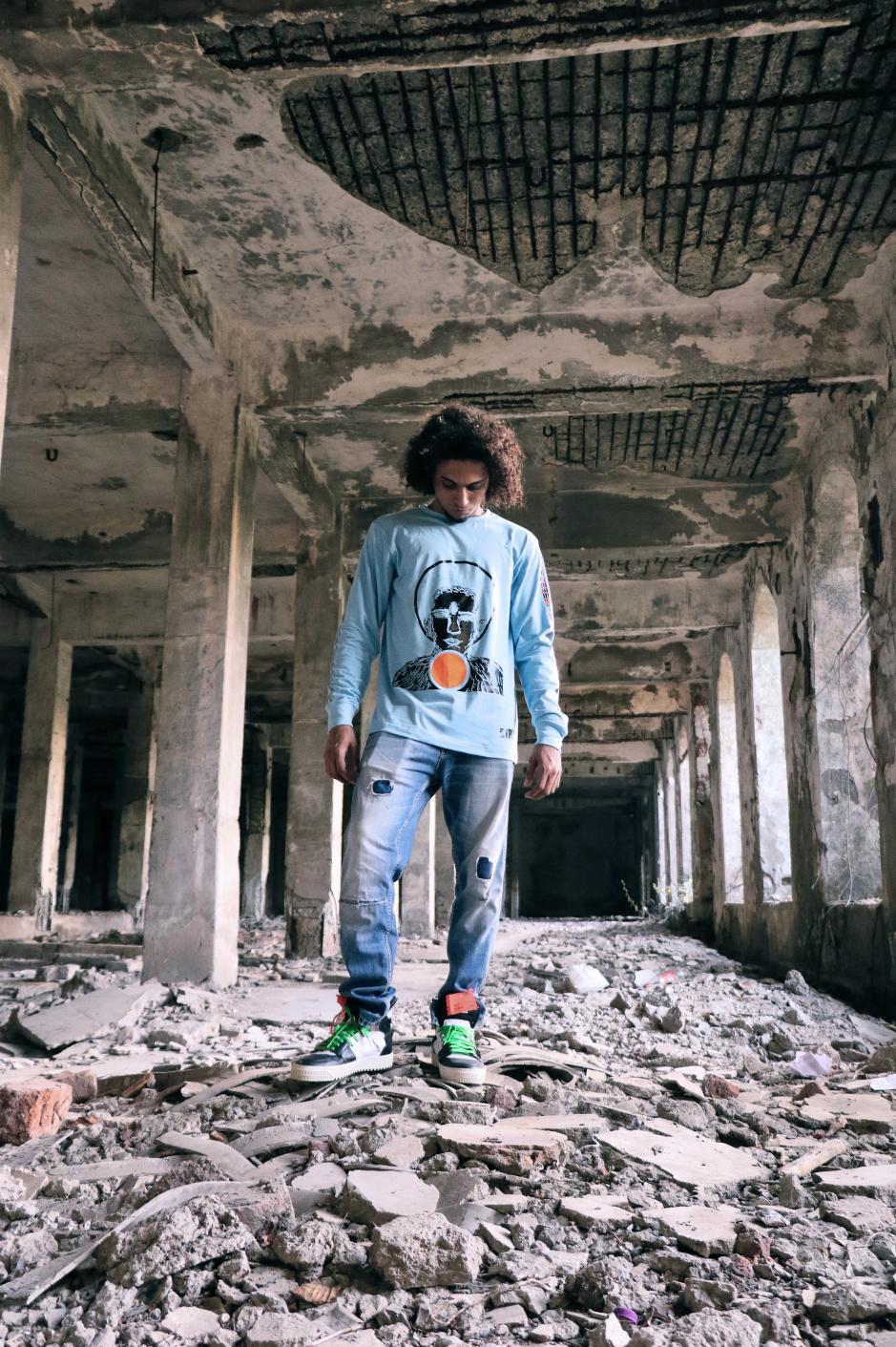 A boy wearing blue high neck full sleeve regular fit T-shirt with a man with three eyes printed on it paired with blue denim and shoes is standing inside a building looking at his feet
