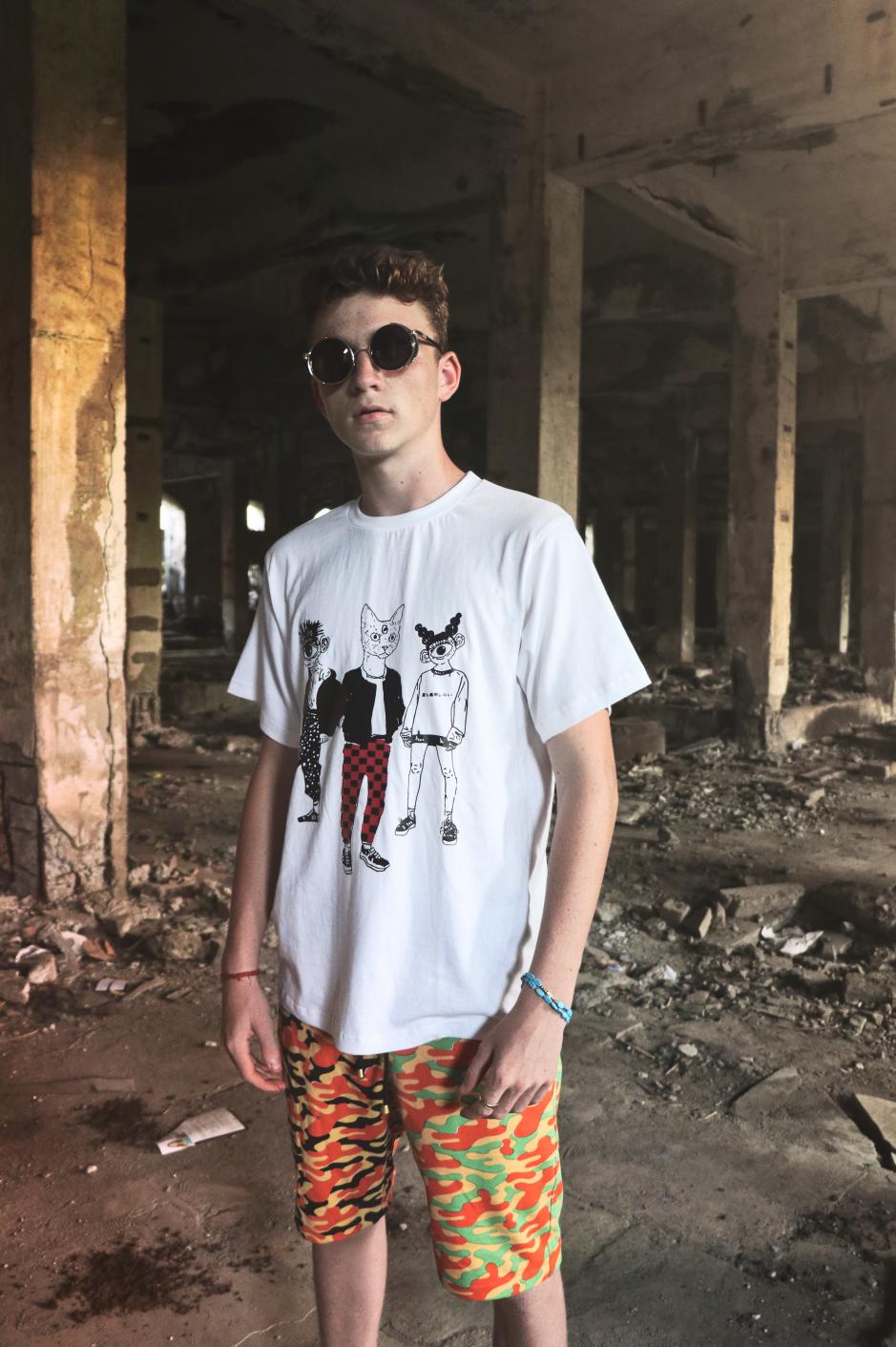 A guy wearing white round neck short sleeve regular fit T-shirt with 3 cartoons printed on it paired with multi color printed shorts is standing inside a building.