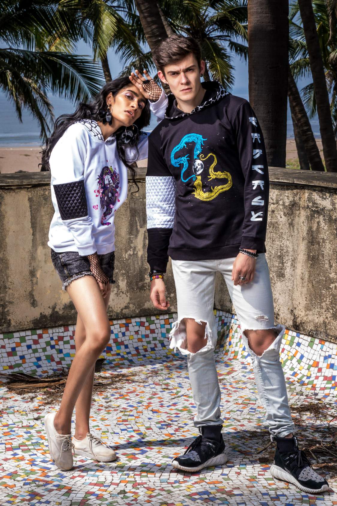 A girl wearing white hoodie with a dancing dragon printed on it with a pair of shorts and white shoes is standing with a guy wearing a black hoodie, knee torn denim and black shoes.