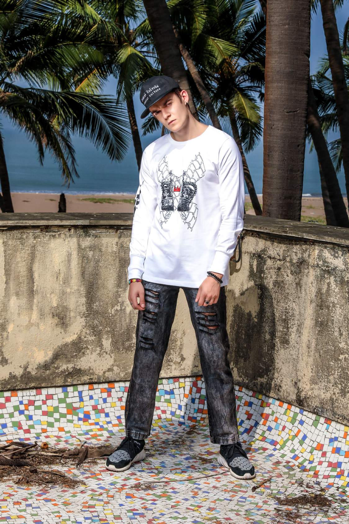 A guy wearing white round neck full sleeve T-shirt with elastic on the wrist, a crown printed in the center paired with faded black denim shoes and a cap is standing on a mosaic floor.